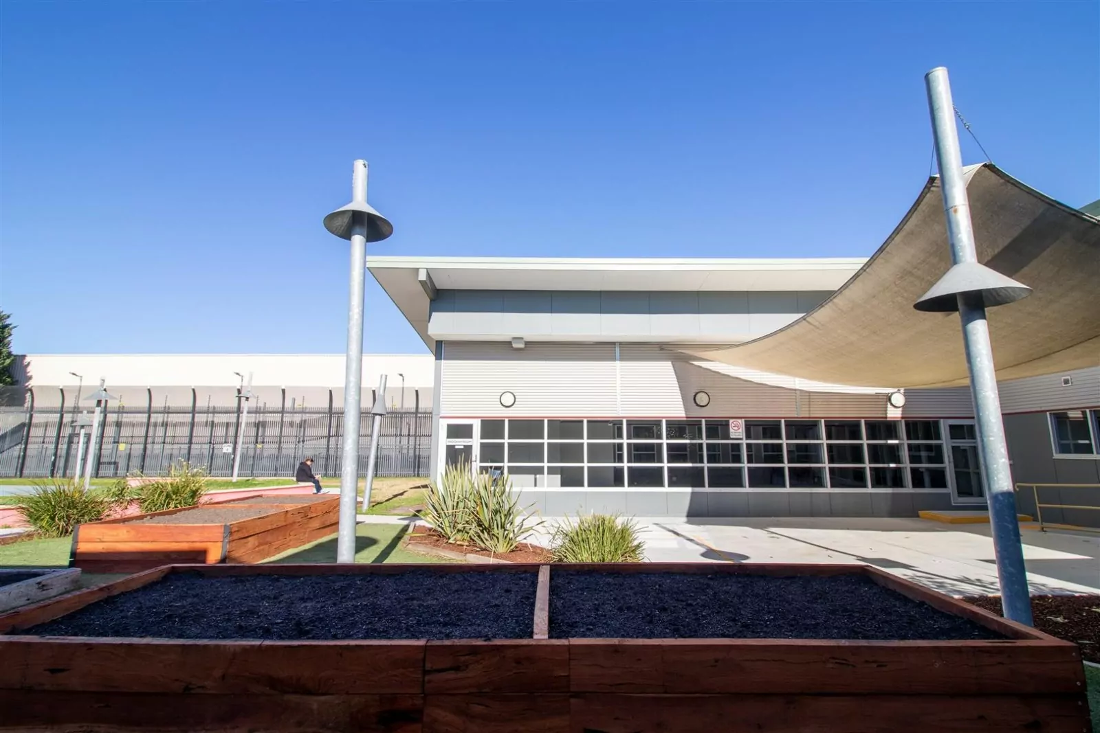 The Maribyrnong Transition Centre providing emergency parolee accommodation for the Department of Justice and Community Safety (DJCS). A person sits in the grass courtyard adjacent to a tall, fenced and secure facility. There are wood paneled flowerbeds with native paints.